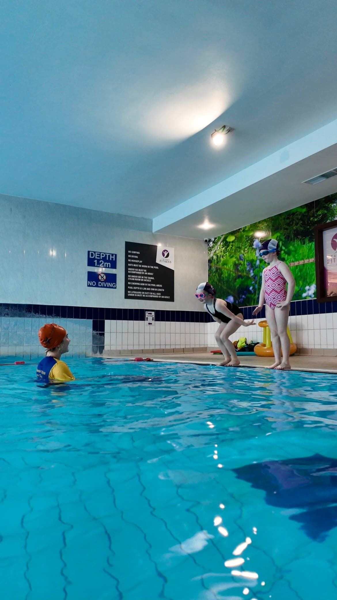Two young swimmers on the poolside preparing to jump in as their teacher waits below