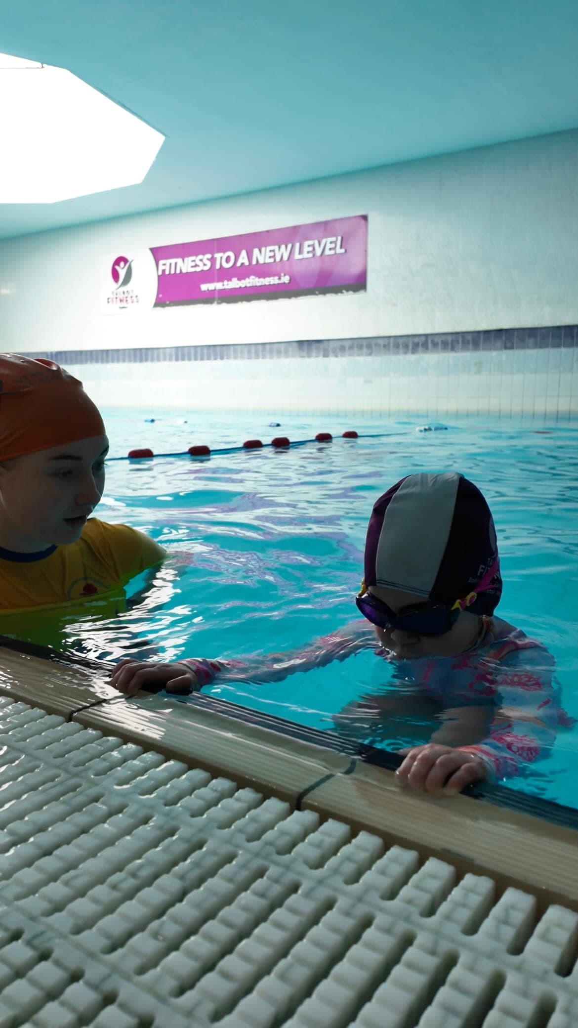 A swim teacher at the wall alongside a young swimmer holding the poolside