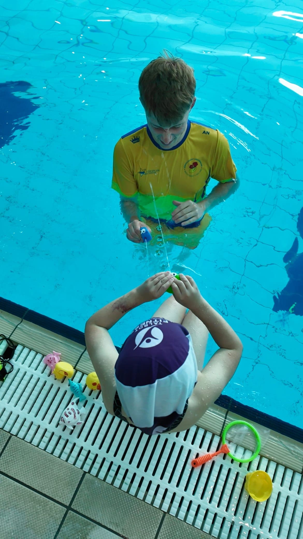 A swim teacher pouring water from a cup over a toddler sitting on the poolside